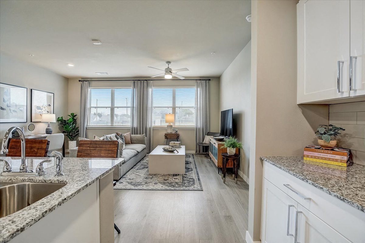 Open living room with large windows, ceiling fan, and kitchen pass-through with granite counters.